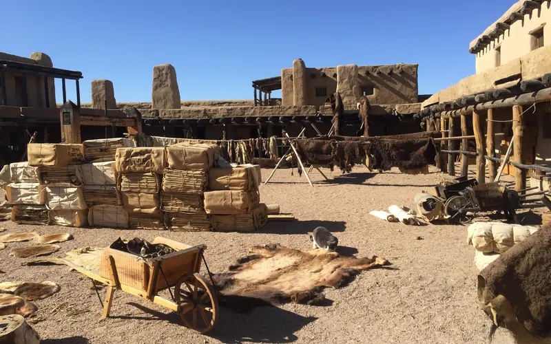Wooden boxes and buffalo furs are scattered around a plaza inside an adobe fortification.