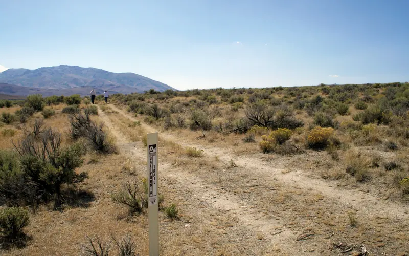 A dirt road passes through sagebrush and a white post with a mountain in the background.