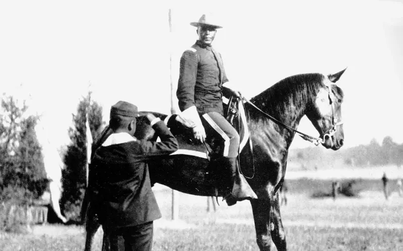 An African American man in a military uniform sitting high on a horse being saluted by a boy