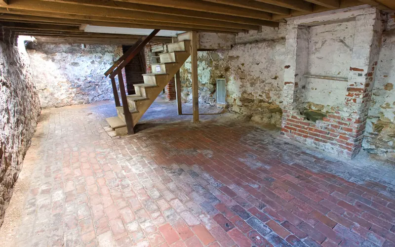 Color photo of the basement with brick floor, false chimney, and wooden staircase.