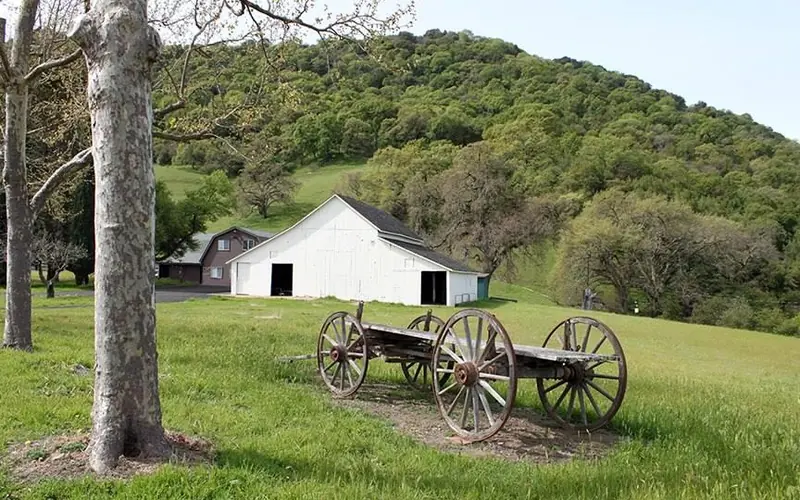 An old barn and an historic wheeled wagon sit in front of a tree-lined hill.