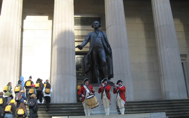 Three people in reenactment clothing hold instruments in front of a stone building and giant statue.