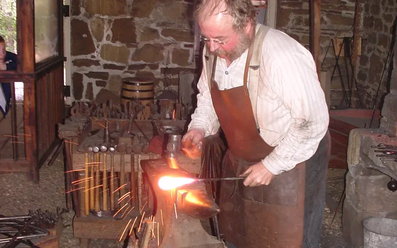 Man in leather apron hammering hot steel in blacksmith shop.