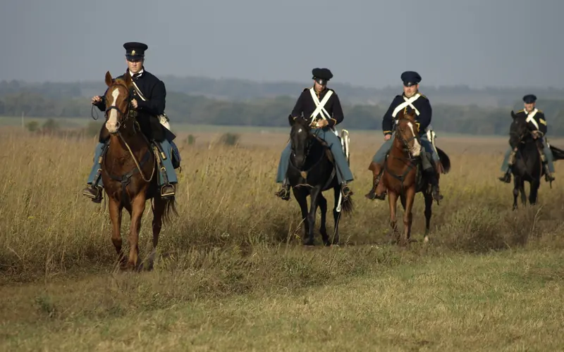 Four soldiers on horseback in a line riding through prairie grass.