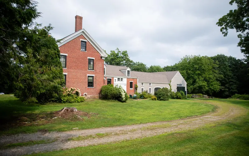 A sandy brown driveway curves through green grass towards the two story-red brick homestead and grey