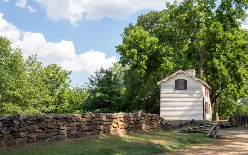 A gravel path bordered by a stone wall leading to a small, two story white house.