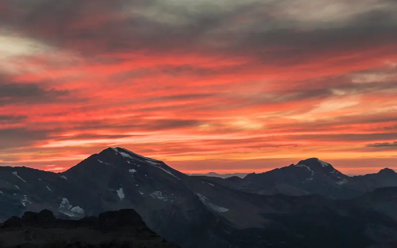 Clouds of orange and red sit above dark-gray mountains; snow dots the mountain peaks.