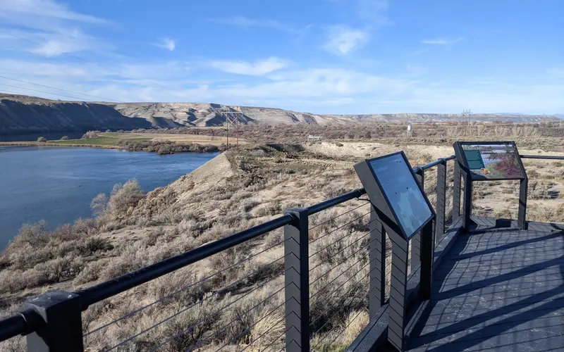 A wooden platform with railing, along with two exhibit signs, overlooks the river and fossil beds.