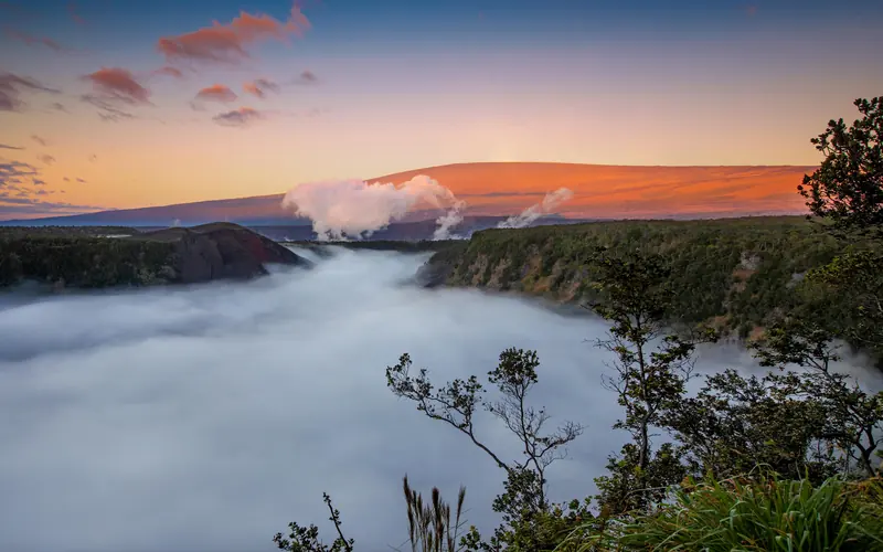 A cloud-filled volcanic crater at sunrise with a mountain rising behind