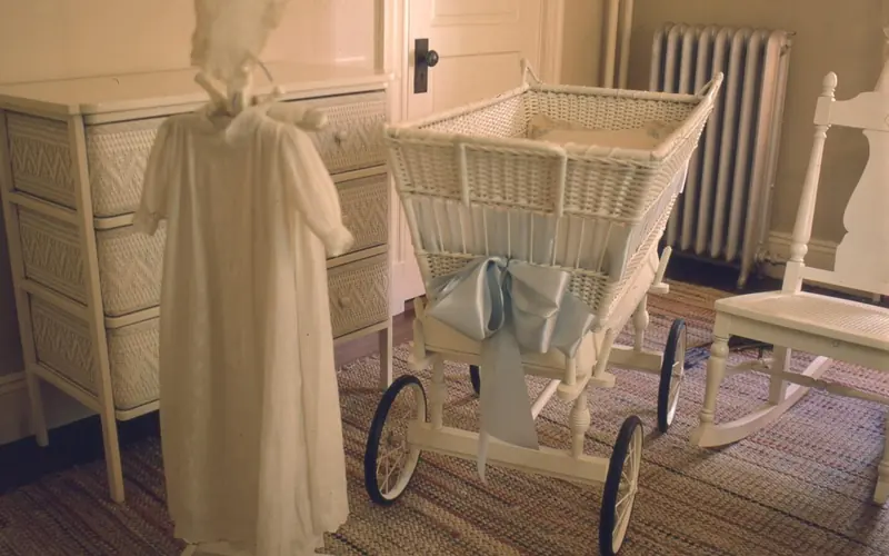 Room with white wicker furniture, including bassinet and rocking chair. Long white baby dress hangs.
