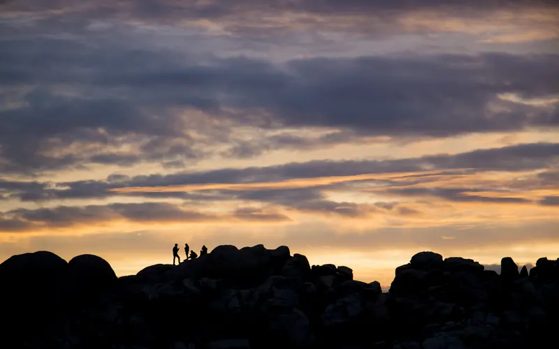 people climbing on boulders are silhouetted against a colorful sunset sky