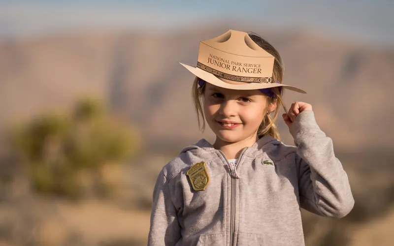 a little girl smiles while wearing a Jr. Ranger hat and badge