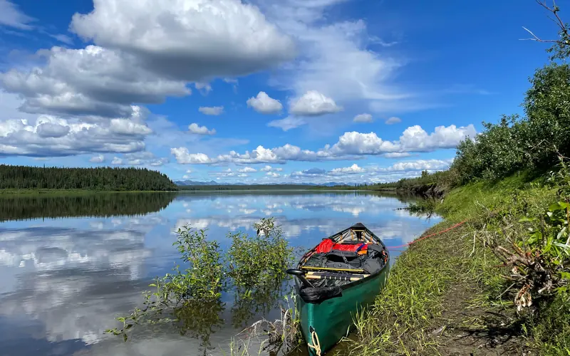 A green pack canoe sits on the bank of the Kobuk River. Clouds are mirrored on the water's surface.
