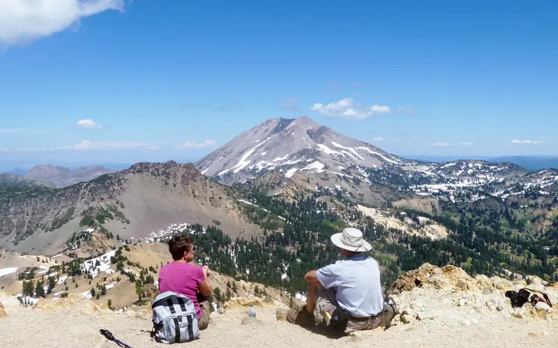 Two hikers sit on a mountain top with a view of multiple peaks, dotted with patches of snow