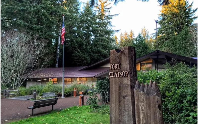 A wooden rectangular sign reading Fort Clatsop eclipsing a wide one story building and flag pole
