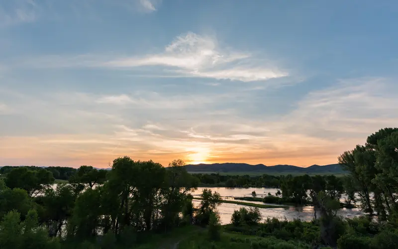 sun dips behind mountains. In the foreground and meandering river catches the light. Leafy trees sur