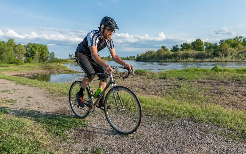 Cyclist biking dirt path along riverfront. Leafy trees and wetlands habitat in distance.