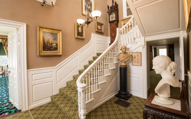 A staircase with white balusters and green carpet. A bust of Washington sits at the turn of stairs.