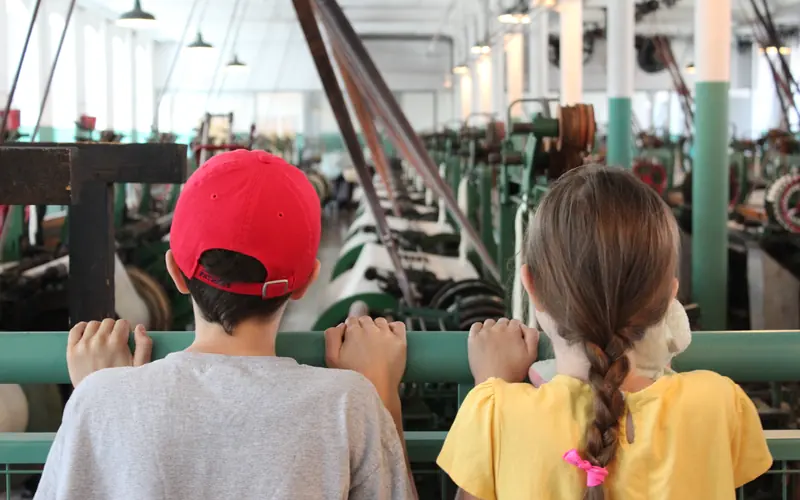 Two young visitors look over the rail at a room full of working looms