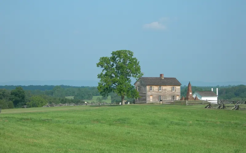 Rustic farmhouse, flanked by shade tree, stone monument, and white outbuilding, on top of a hill.
