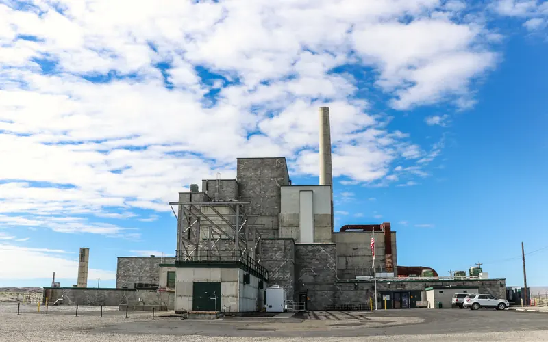 Gray factory building like stacked boxes with single taller smokestack beneath cloudy blue skies.