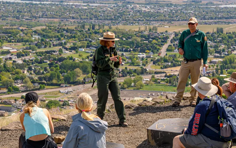 Uniformed park ranger talks animatedly to visitors on a hillside overlooking a sprawling urban area.