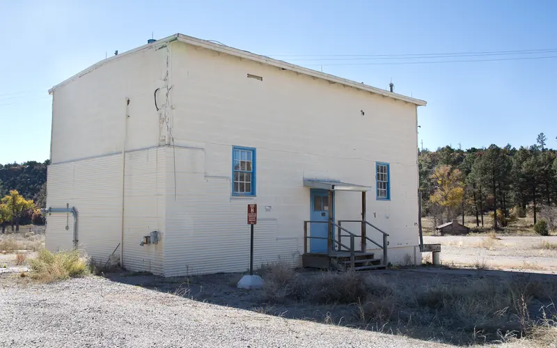 Two-story white house with two small windows, a single door, and flat roof stands in a gravel lot.