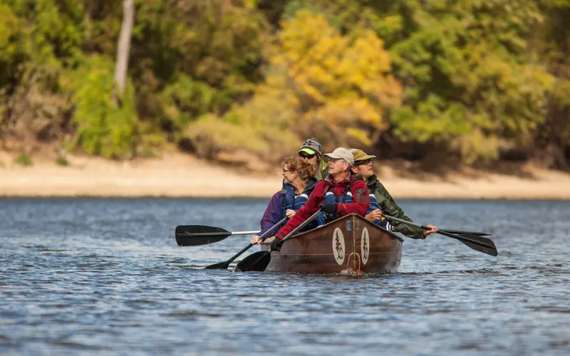 A large canoe filled with paddlers on the river surrounded by autumnal color.