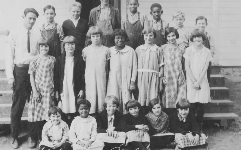 Twenty children of mixed ages and genders posing for the camera in front of a schoolhouse.