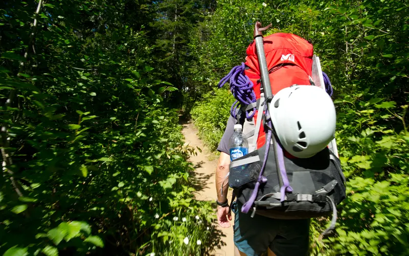 Person with backpacking and climbing gear hikes on a trail.