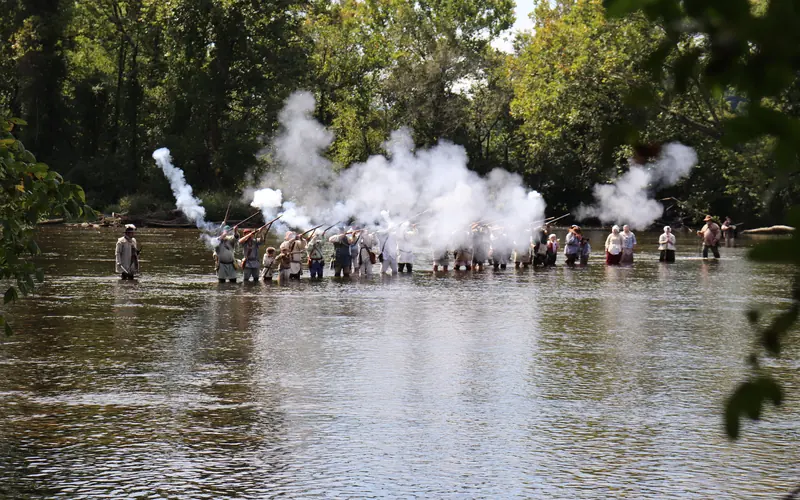 A large group of living historians stand in a river and fire muskets, with a cloud of smoke rising.