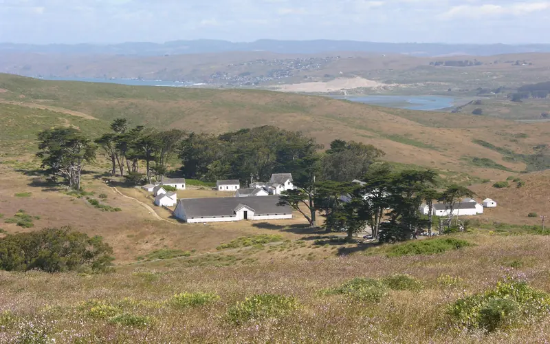 A historic dairy ranch composed of white-painted buildings surrounded by dry grass and a few trees.