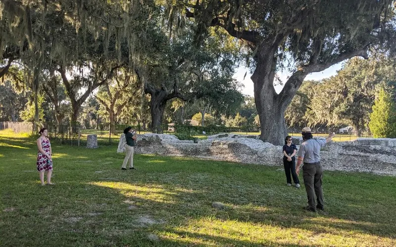 A Park Ranger points at the ruins of a tabby wall with three people looking on