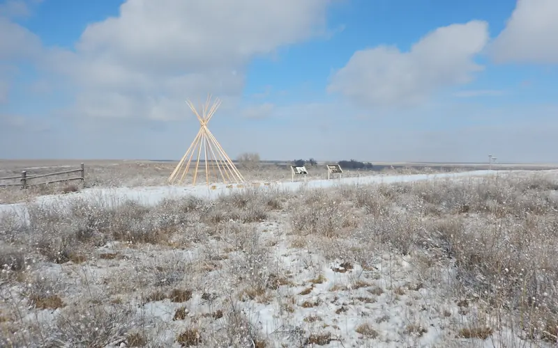 A lone tipi frame and two wayside signs stand in a snow-covered landscape