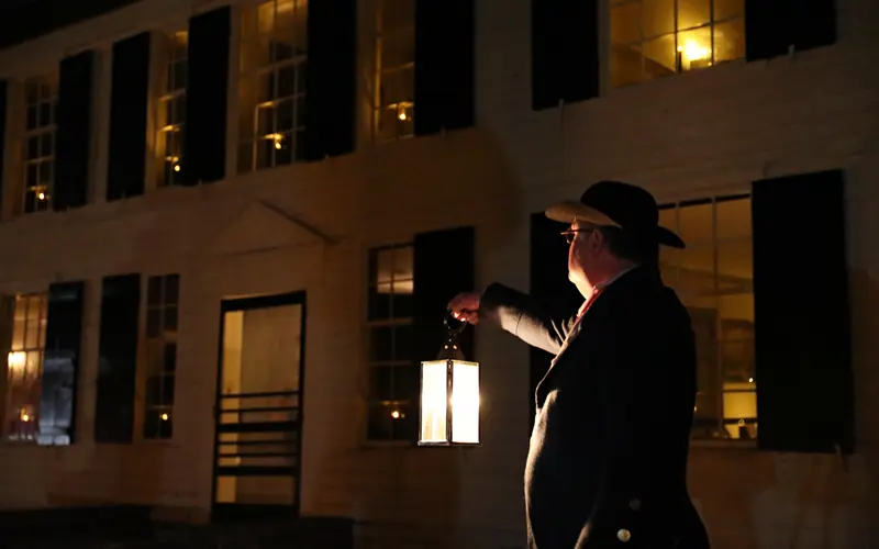 A man in 18th-century style clothing holds a candlelit lantern in front of a 2-story yellow house.