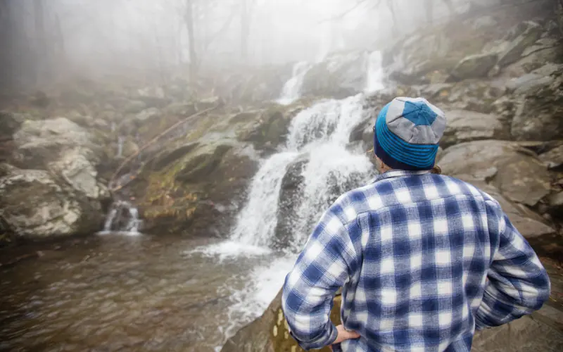 A man stands with his back facing us, looking at a waterfall.