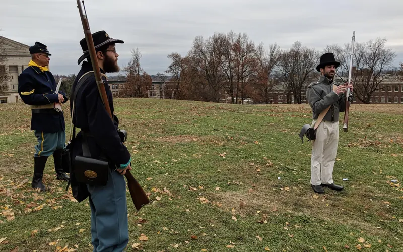 Three Park Volunteers, holding historic firearms, on the grounds of the Armory against a cloudy sky.