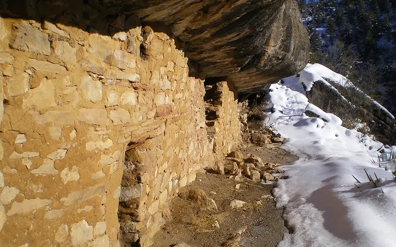 warm sunlight illuminates a cliff dwelling wall beside a snowy trail