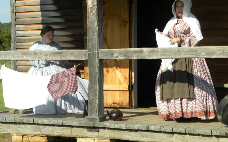 Women in reproduction historic dress sit and stand on porch of old cabin