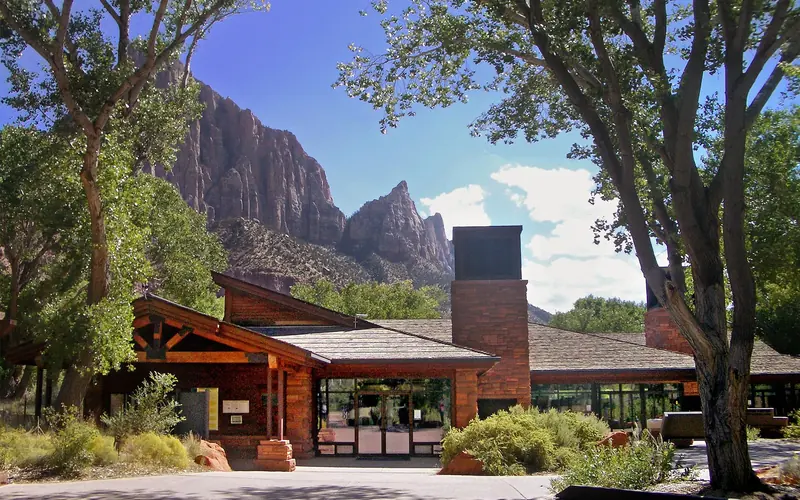A sandstone and glass building surrounded by tall trees with a mountain behind it.